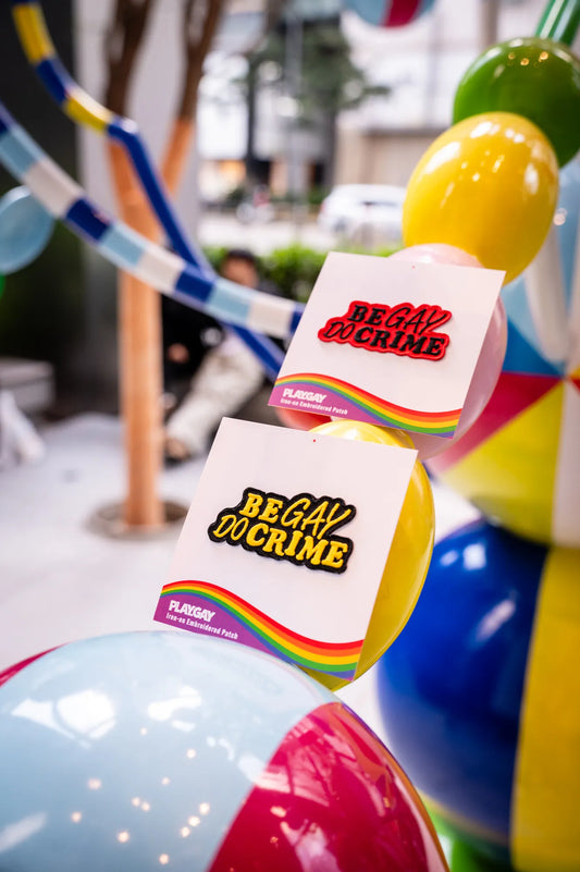 Two “Be Gay Do Crime” iron-on patches in red and yellow, displayed on PlayGay cards in front of colorful balls.