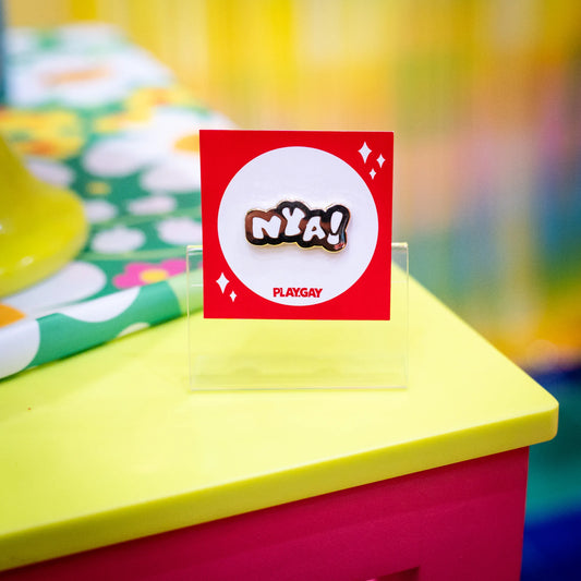 An enamel pin with the text "Nya!" in white and gold design, attached to a red PlayGay backing card, displayed in a colorful and fluffy anime-themed setup.