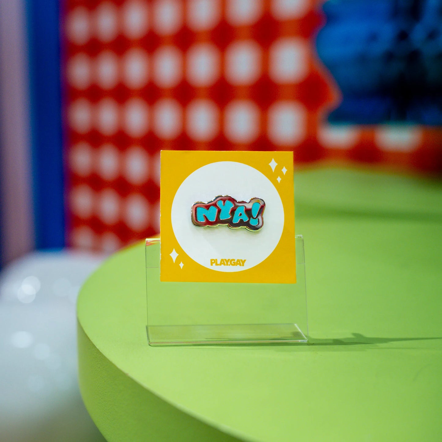 An enamel pin with the text "Nya!" in blue and silver color, attached to a yellow PlayGay backing card, displayed in a colorful and fluffy anime-themed setup.