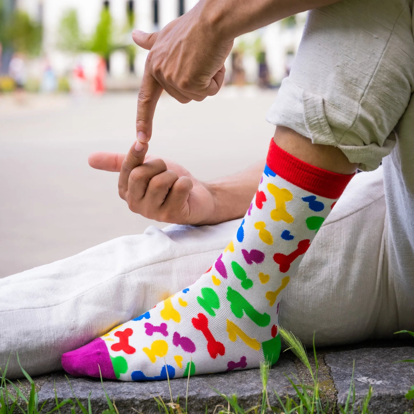 Close-up of person wearing white socks with rainbow-colored toy shapes and red cuff, sitting on pavement with suggestive hand gesture.