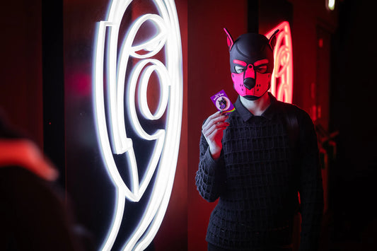 A puppy in a red hood holding up a puppy enamel pin in purple colors on a rainbow PlayGay backing card with the words "Who is a good boy?