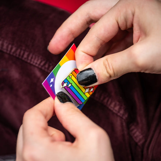 A person mounting an Enamel in the shape of the rainbow LGBTQ+ pride flag, decorated with white sparkles on a rainbow-themed PlayGay backing card.
