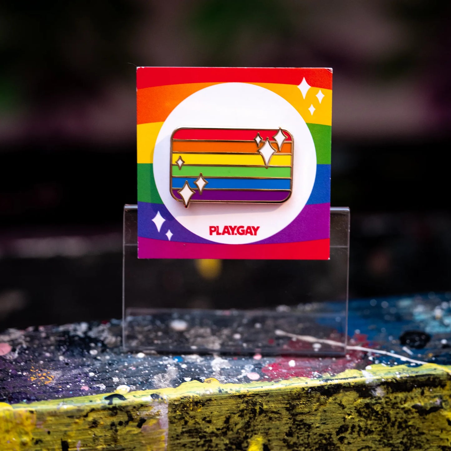 Enamel pin in the shape of the rainbow LGBTQ+ pride flag, decorated with white sparkles and mounted on a rainbow-themed PlayGay backing card with a blue & yellow background.