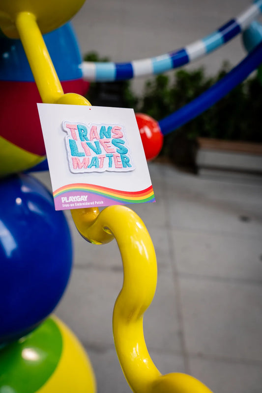 Pastel-colored patch reading Trans Lives Matter in the colors of the trans flag, attached to a PlayGay backing card.