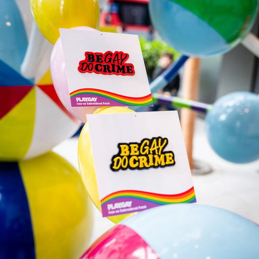 Two “Be Gay Do Crime” iron-on patches in red and yellow, displayed on PlayGay cards in front of colorful balls.