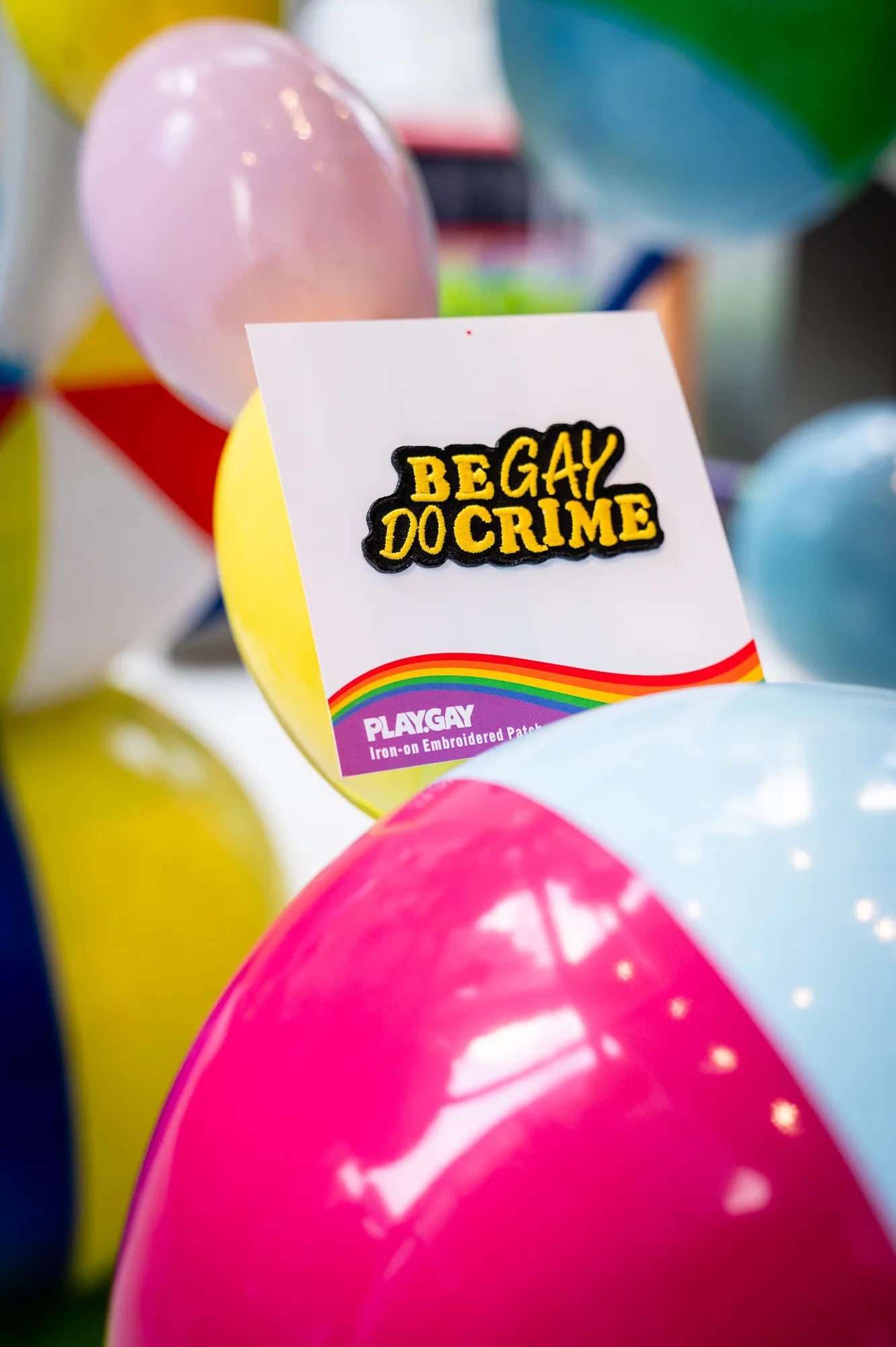 A "Be Gay Do Crime” iron-on patch in yellow displayed on PlayGay cards in front of colorful balls.