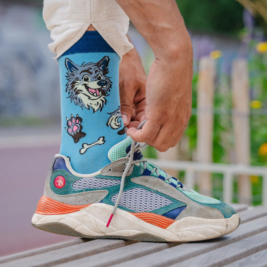 Close-up of blue socks with a cartoon wolf, paw print, and bones, worn with colorful sneakers while tying shoelaces on a bench.