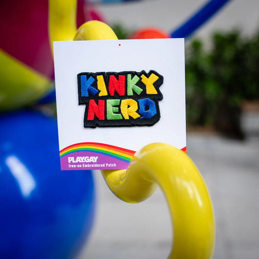 Colorful embroidered patch reading Kinky Nerd in rainbow letters attached to a PlayGay card with rainbow branding, shown in a playful balloon installation.