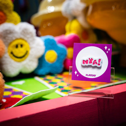 An enamel pin with the text "Nya!" in white and pink design, attached to a purple PlayGay backing card, displayed in a colorful and fluffy anime-themed setup.