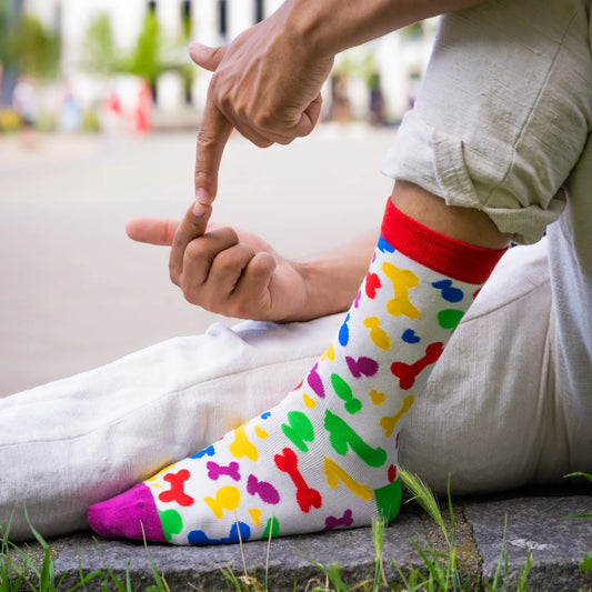 Close-up of person wearing white socks with rainbow-colored toy shapes and red cuff, sitting on pavement with suggestive hand gesture.