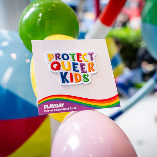 Rainbow embroidered “Protect Queer Kids” patch by PlayGay, photographed in a playful, colorful environment with balloons in the background.