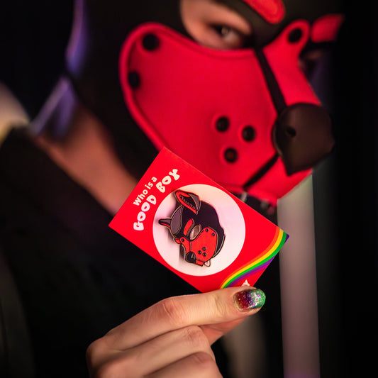 A puppy in a red hood holding up a puppy enamel pin in red colours on a rainbow PlayGay backing card with the words "Who is a good boy?