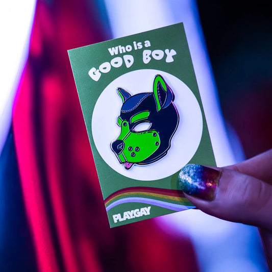 A guy with rainbow nails holding up a puppy enamel pin in green colors on a rainbow PlayGay backing card with the words "Who is a good boy?