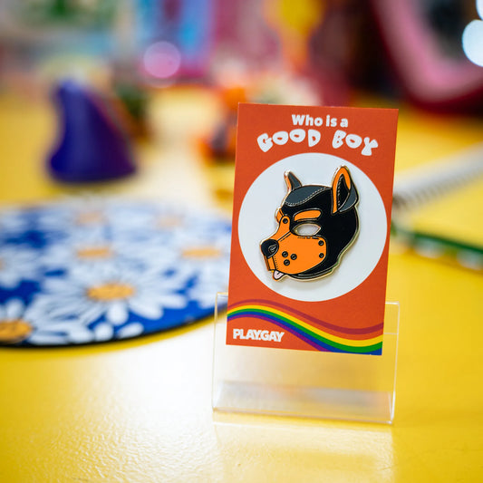 A puppy enamel pin in orange colors on a rainbow PlayGay backing card with the words "Who is a good boy? standing in a funky colored room.