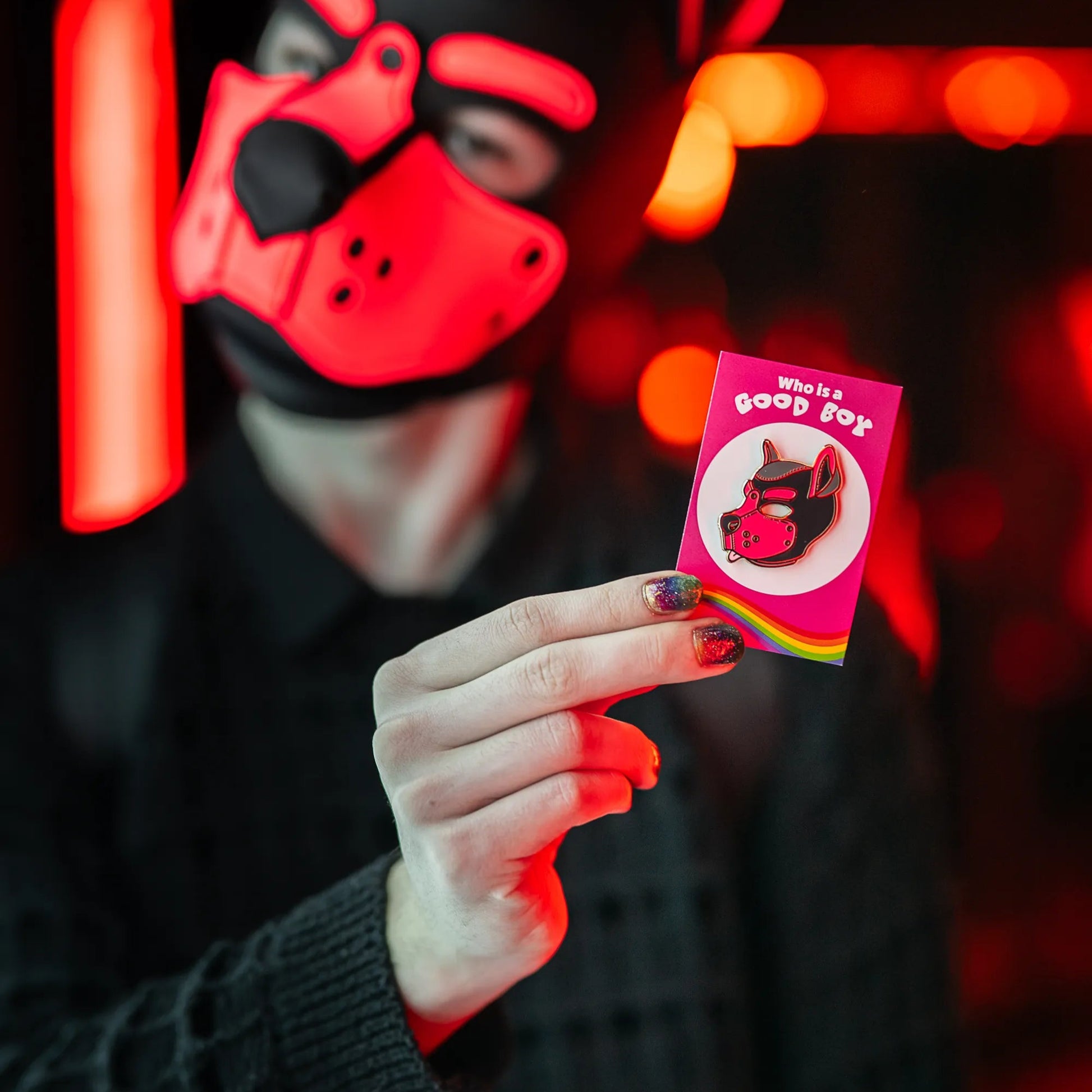 A puppy in a red hood holding up a puppy enamel pin in pink colors on a rainbow PlayGay backing card with the words "Who is a good boy?