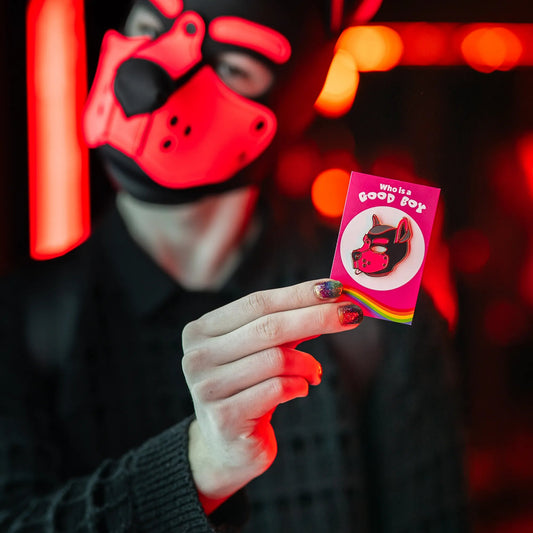 A puppy in a red hood holding up a puppy enamel pin in pink colors on a rainbow PlayGay backing card with the words "Who is a good boy?