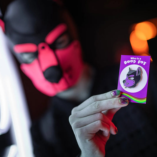 A puppy in a red hood holding up a puppy enamel pin in purple colors on a rainbow PlayGay backing card with the words "Who is a good boy?