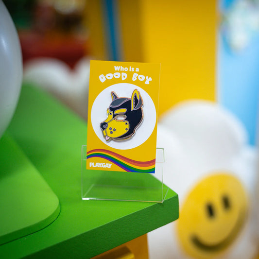 A puppy enamel pin in yellow colors on a rainbow PlayGay backing card with the words "Who is a good boy? standing in a funky colored room.
