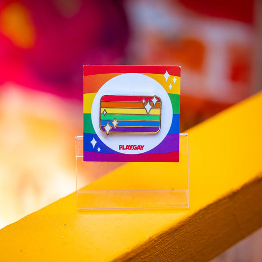 Enamel pin in the shape of the rainbow LGBTQ+ pride flag, decorated with white sparkles and mounted on a rainbow-themed PlayGay backing card.