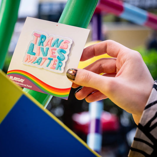 Pastel-colored patch reading Trans Lives Matter in the colors of the trans flag, attached to a PlayGay backing card and held by a person with glittery nails.
