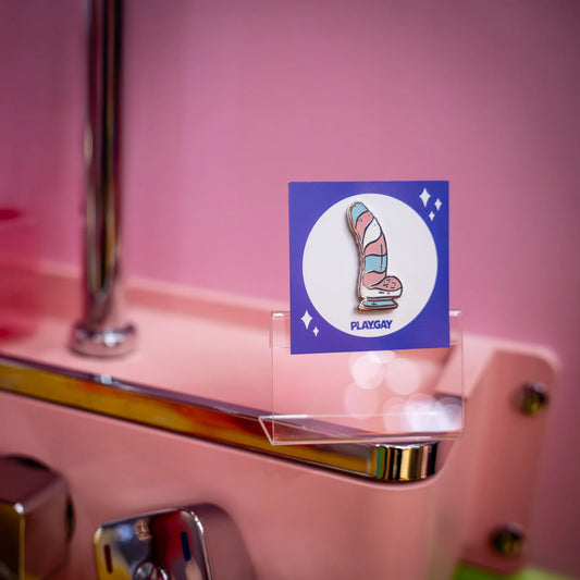 Hard enamel pin featuring a pastel dildo design in the colors of the trans pride flag, mounted on a purple PlayGay card, displayed in a bathroom setup.