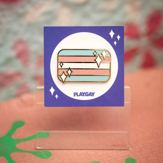 Trans pride flag enamel pin with pastel blue, pink, and white horizontal stripes, decorated with white sparkles, mounted on a PlayGay card.